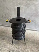 Fogão a lenha preto com alças amarelas, design artesanal, apoiado em três pernas, sobre piso cinza