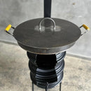 Fogão a lenha artesanal com tampa metálica e alças amarelas em ambiente interno cinza