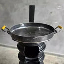 Chapa de ferro redonda com duas alças amarelas sobre suporte preto, fundo cinza