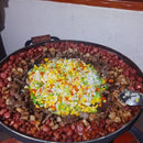 Panela grande com legumes picados no centro cercados por carnes variadas assando em fogão