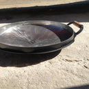 Frigideira redonda de ferro com alça de madeira apoiada sobre chão de cimento ao ar livre