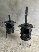 Duas fogueiras negras de metal com suporte, alças amarelas e chaminés em piso de concreto e parede cinza